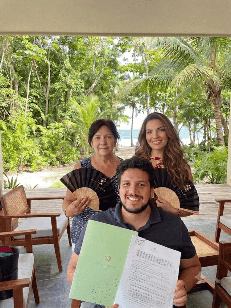 Tres personas sonriendo en una terraza con vista al mar el hombre sostiene una carpeta con un documento impreso